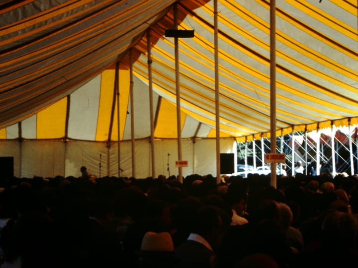 inside of a blue and yellow striped tent, a seated crowd is gathered