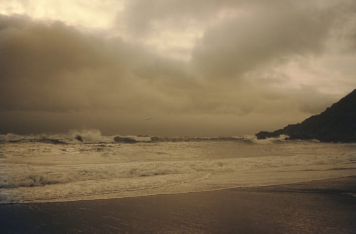 waves crashing on a beach with a headland to the right and cloudy sky